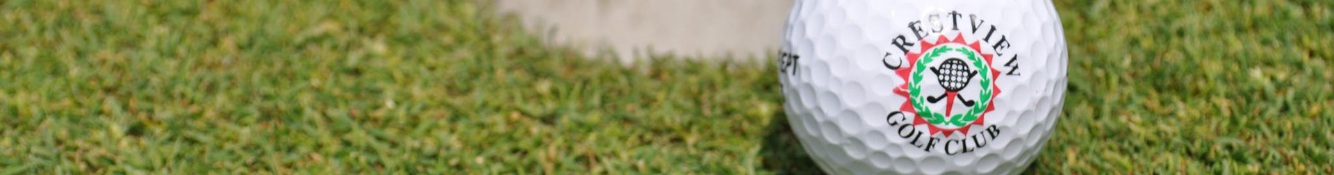 A close-up of a Crestview Golf Club logo on a golf ball lying on lush green grass, with a sunny golf course in the background.